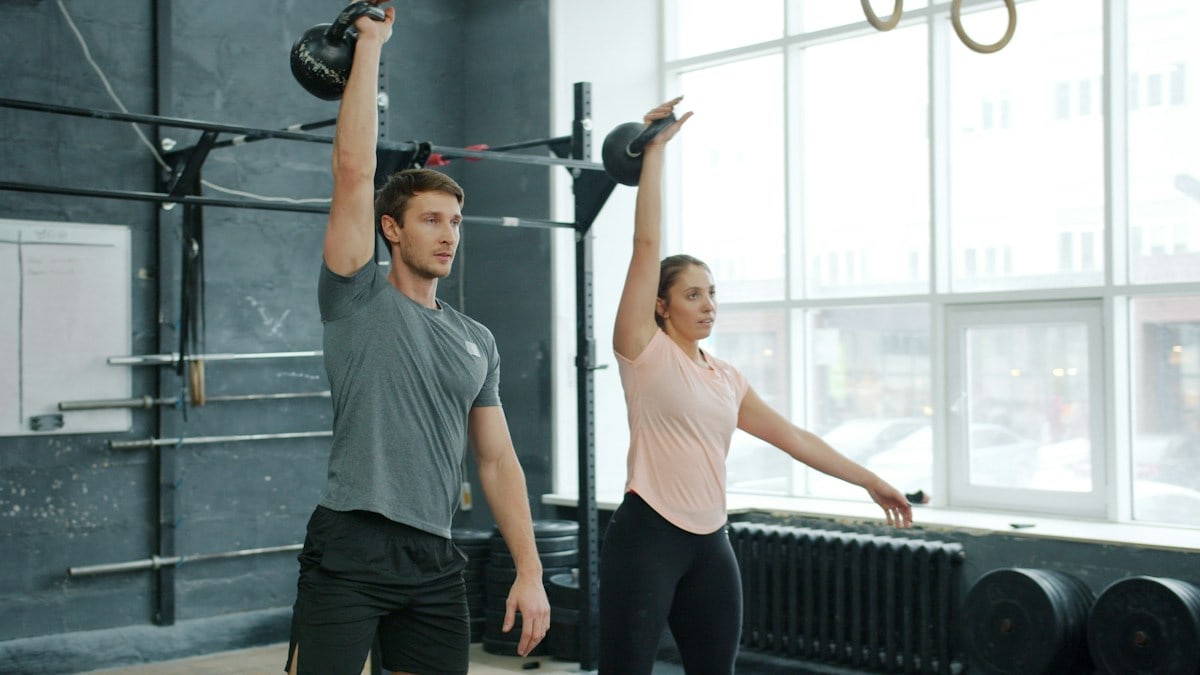 Young couple man and woman are training in gym weightlifting squatting with kettlebells together concentrated on bodybuilding exercises indoors.